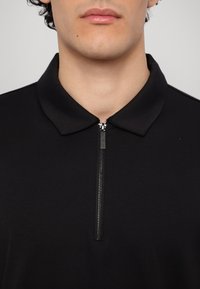 Svart polo-skjorte med glidelås krage, laget av et glatt stoff. Den har en kort glidelås ved halsen og en subtil glans.
