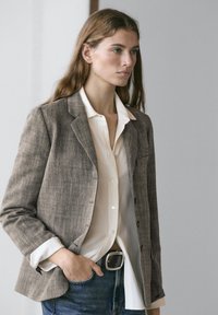 Grå linnedblazer med strukturerede skuldre, enkelt knaplukning og to forlommer, kombineret med en hvid skjorte med krave og blå jeans.