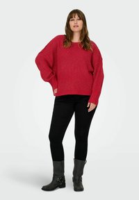 Maglione rosso a maglia con maniche larghe, indossato sopra jeans skinny neri e stivali alla caviglia grigi con fibbie. Tessuto morbido, vestibilità rilassata.