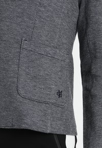 Detailní záběr texturovaného šedého saka s jemným černým vyšitým logem v dolním rohu kapsy, zobrazující část rukávu a černé kalhoty.