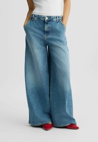 Jeans a gamba larga in denim azzurro chiaro con vita alta, dotati di tasche anteriori, chiusura con bottone e cuciture a contrasto. Abbinati a scarpe rosse.