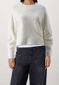 Vaaleanharmaa lyhyt neulepaita, jossa on pitkät hihat ja pyöreä pääntie, kerroksittain valkoisen paidan päällä, yhdistettynä tummiin farkkuihin, joissa on näkyvät saumat.