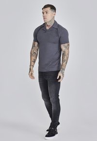 Polo de color gris oscuro con cuello puntiagudo, mangas cortas y un logo sutil. Combinado con jeans negros de corte slim y zapatillas negras con suelas blancas.