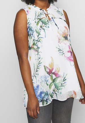 Kvinde iført ærmeløs hvid bluse med flerfarvet blomstermønster og bindedetalje ved halsudskæringen, kombineret med mørkegrå bukser.