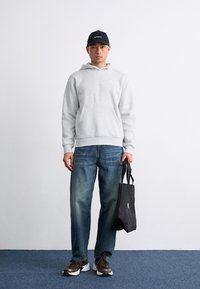 Sudadera gris con capucha y bolsillo frontal, jeans azules, zapatillas oscuras y una gorra negra. Sosteniendo una pequeña bolsa tote negra. Fondo neutro.