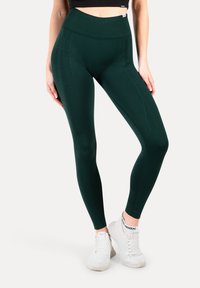 Donkergroene leggings met een hoge tailleband, een gladde textuur en subtiele branding aan de zijkant, gecombineerd met witte sportschoenen.