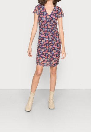 Vrouw draagt een nauwsluitende, korte mouwen, geplooide bloemenjurk in marineblauw met roze en oranje bloemen, gecombineerd met beige enkellaarsjes.