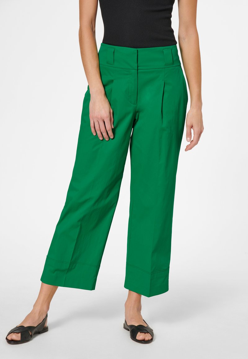 Groene wijde broek gemaakt van een gladde, lichte stof. Kenmerkt zich door voorplooien, een hoge taille en een cropped zoom. Zwarte sandalen worden gedragen.