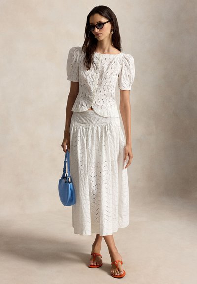 Γυναίκα που φοράει άσπρο πουκάμισο με μανίκια-φουσκωμένα από κεντήματα και μεμονωμένη φούστα, κρατώντας μπλε τσάντα, με σκούρα γυαλιά ηλίου και πορτοκαλί σανδάλια σε μπεζ φόντο.