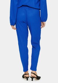 Modré, lehké texturované kalhoty s úzkým střihem a elastickým pasem, doplněné černými sandály na otevřenou špičku.