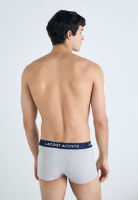 Graue Baumwoll-Boxershorts mit einem marineblauen Bund, auf dem der Schriftzug "LACOSTE" steht. Das Modell steht mit dem Rücken zum Betrachter.