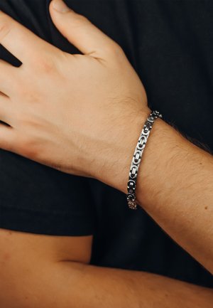Silver metal bracelet with interlocking chain links and black accents, worn on a wrist with a natural skin tone, against a black shirt.