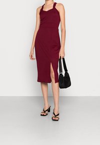 Robe midi ajustée bordeaux avec un décolleté carré, une texture côtelée et une fente latérale ; associée à des sandales noires à lanières et un sac bandoulière noir.