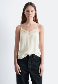 Cremfarbene Satin-Cami mit Spitze am Ausschnitt, ausgestattet mit dünnen verstellbaren Trägern, kombiniert mit hochtaillierten schwarzen Jeans.