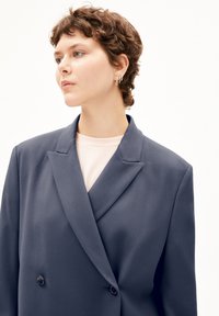 Blazer doppiopetto in tessuto blu scuro con una superficie liscia. Presenta un ampio colletto, rever a punta e due bottoni neri.