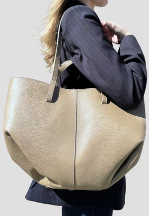 Donna bionda in blazer scuro che porta una grande borsa tote in pelle beige testurizzata sulla spalla, su sfondo semplice.