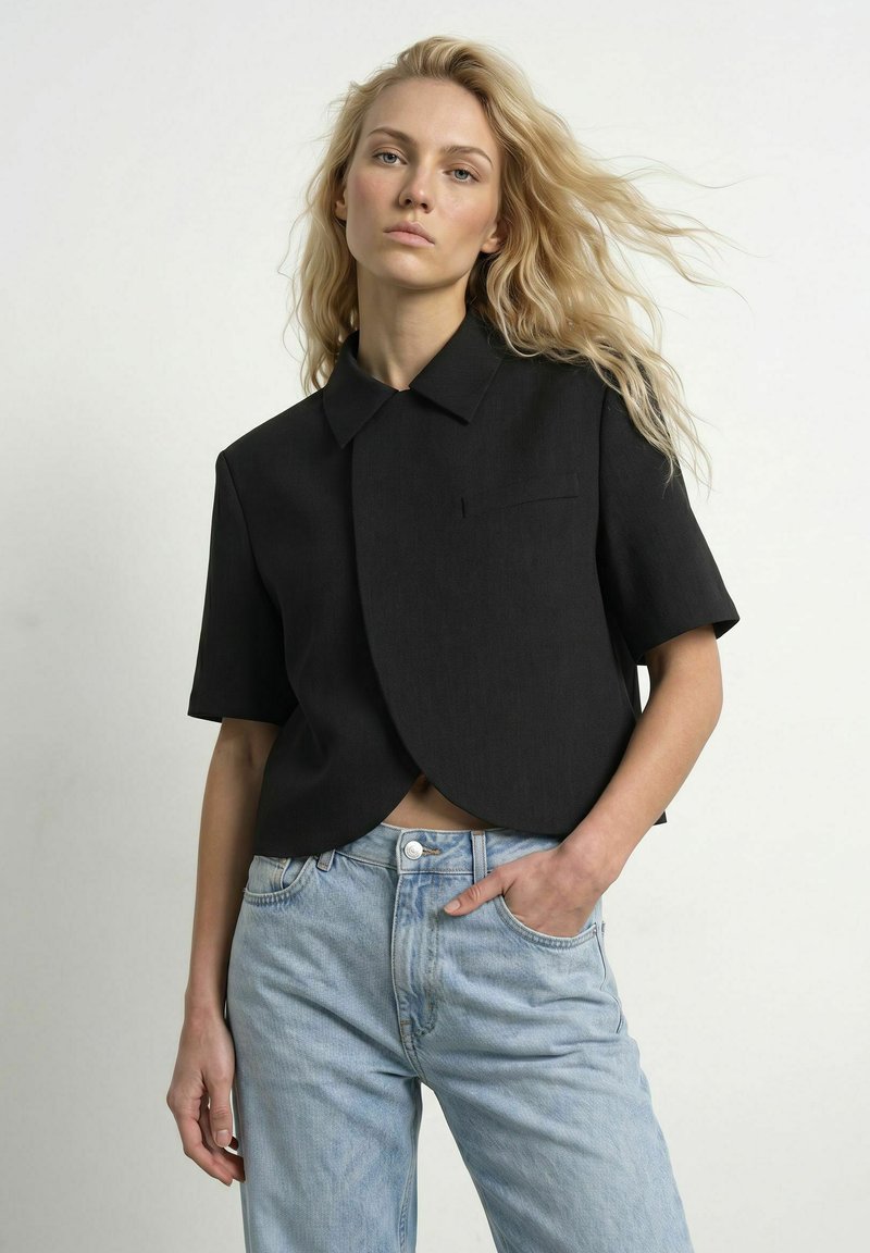 Femme blonde aux cheveux ondulés portant un blazer noir à manches courtes et un jean taille haute bleu clair, debout avec une main dans la poche devant un mur blanc.