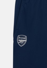 Jūras zilas audums ar baltu „Arsenal” futbola kluba logotipu, kurā attēlots lielgabals un teksts apakšējā kabatas daļā.
