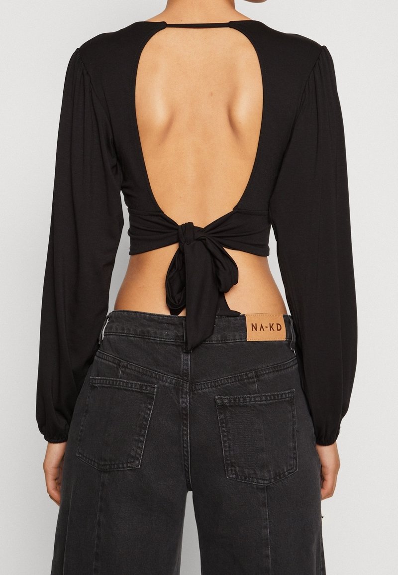 Svart långärmad topp med öppen ryggdesign, knuten vid midjan. Mjukt tyg, slät textur. Kombination med högmidjade svarta denimjeans.