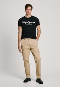 Crna majica s bijelim logotipom "Pepe Jeans London", bež cargo hlače s džepovima i crne cipele. Jednostavan, ležeran outfit.