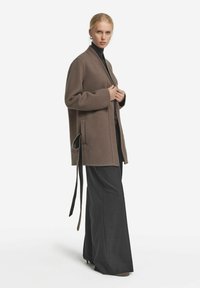 Brun omslagsjacka i mjukt tyg med långa ärmar och två framfickor. Har en knytning i midjan. Kombinerad med svarta vida byxor.