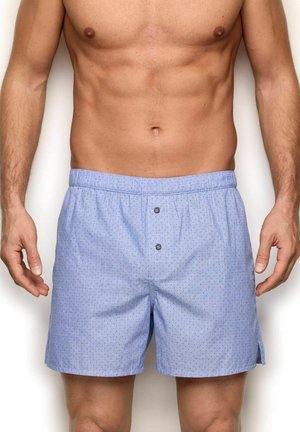 Męski tors ubrany w jasnoniebieskie bokserki w wzory z dwoma guzikami z przodu, stojący na jednolitym białym tle.