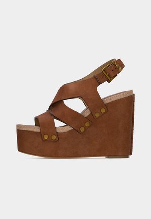 Brun ruskinds kilehælssandal med tre remme, messingnitter og justerbar ankelspænde på en platformsål mod en ensfarvet baggrund.