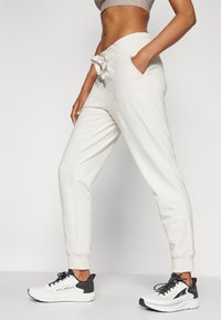 Lys beige joggebukser med snor i livet, sidelommer og ribbede ankler, parret med svart og hvit løpesko.