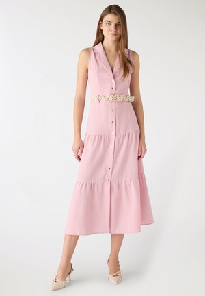 Giovane donna che indossa un abito midi senza maniche rosa con bottoni, cintura intrecciata e tacchi beige a punta, in piedi contro uno sfondo semplice.