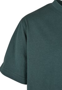 Prim-plan al unui tricou verde cu mâneci scurte, care arată mâneca, cusătura umărului și decolteul rotund pe un fundal alb.