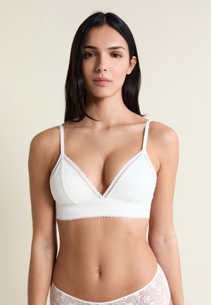 Kobieta o długich ciemnych włosach, ubrana w biały koronkowy stanik typu bralette i pasujące koronkowe majtki na neutralnym tle.