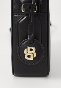 Borsa in pelle nera con un accento circolare in metallo dorato con logo, superficie testurizzata e dettaglio della tracolla, che mette in evidenza il suo design strutturato.