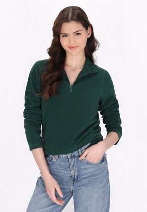Giovane donna con lunghi capelli castani, che indossa un pile verde scuro con zip a un quarto e jeans azzurri chiari, in posa con una mano in tasca.