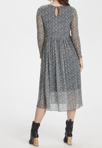 Vestido floral con mangas largas transparentes, detalle de abertura en la espalda y cintura fruncida. Los colores incluyen un patrón floral en azul, amarillo y marrón.