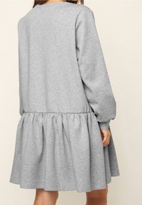 Femme portant une robe longue à manches longues et ample, de couleur gris clair avec une jupe froncée, vue de dos sur fond uni.