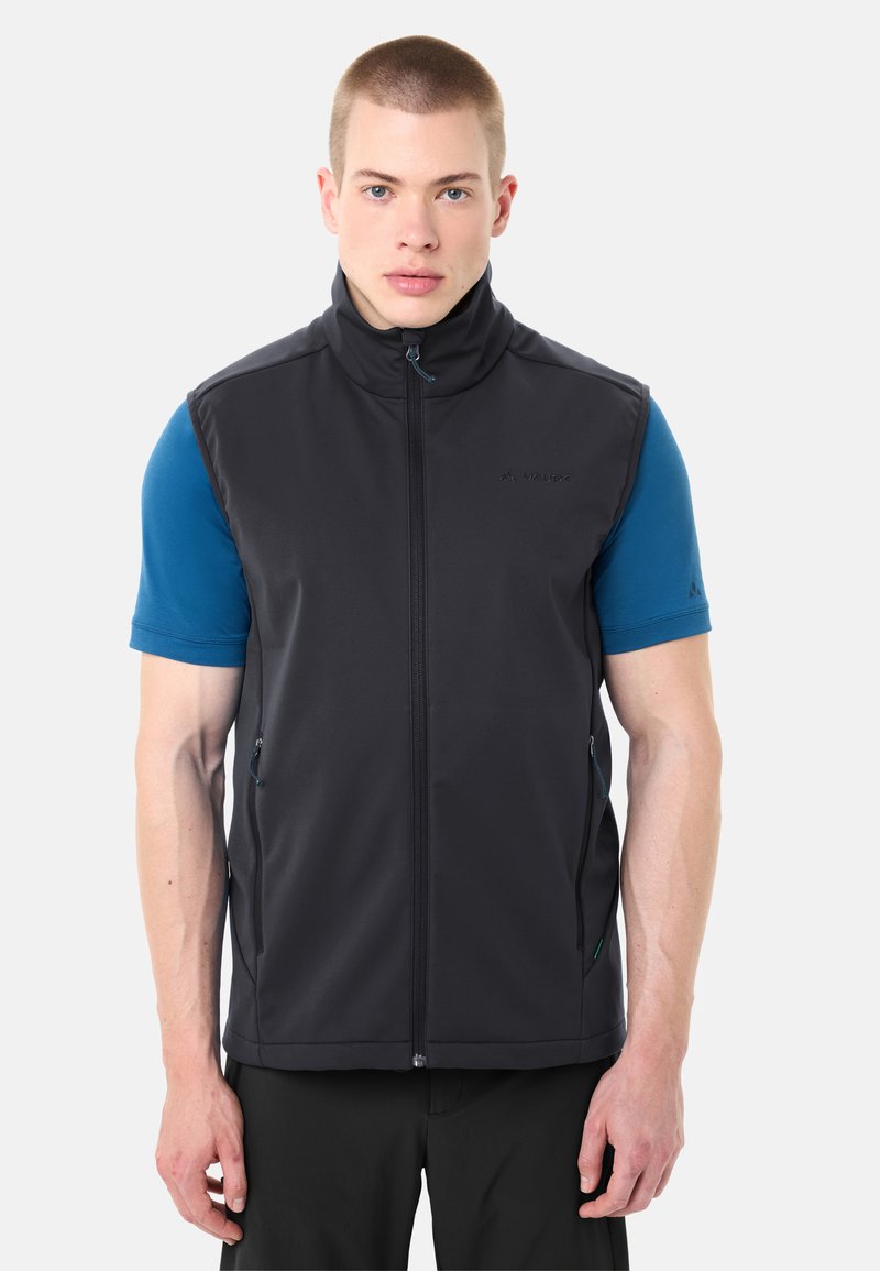 Junger Mann trägt eine schwarze ärmellose Weste mit Reißverschluss über einem blauen kurzärmeligen Hemd, steht vor einem weißen Hintergrund.