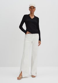 Zwarte top met lange mouwen, een V-hals en een golfrand, gecombineerd met hooggesneden witte wijde broek met zijsplitten en lichte sandalen.