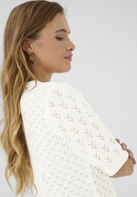 Pull en tricot blanc à manches courtes, présentant un design ajouré et un motif croisé subtil. Texture lisse, encolure ronde.