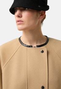 Beige uldblandet frakke med rund hals, sort læderkant og trykknapper, der har en glat tekstur og raglanærmer.