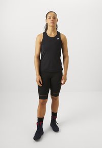 Čierny športový tielko s okrúhlym výstrihom, v kombinácii s čiernymi šortkami, ktoré majú priesvitné panely a textilné pásy. Nosí sa s tmavými športovými topánkami.