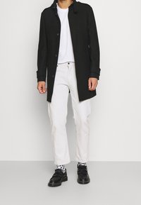 Zwarte wollen jas met knoopsluiting en staande kraag, gedragen over een witte t-shirt en lichte broeken, gecombineerd met zwarte schoenen en gemusterde sokken.