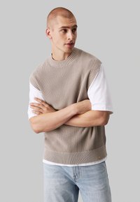 Maglione lavorato a maglia beige senza maniche con un motivo testurizzato, scollatura tonda e orlo a coste, indossato sopra una t-shirt bianca e jeans azzurri chiari.
