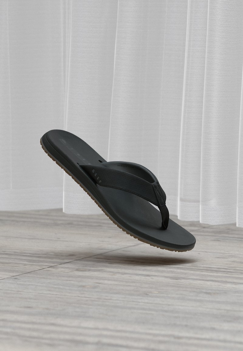 Μαύρο σανδάλι-σαγιονάρα με υφασμένο λουράκι και καφέ σόλα, αιωρούμενο πάνω από ξύλινο πάτωμα μπροστά από λευκές κουρτίνες.