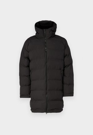 Fekete puffermellény kapucnival, elöl cipzárral, bordázott mandzsettákkal és steppelt kialakítással, fehér ing és fekete nadrág felett viselve.