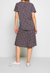 Robe florale noire à manches courtes, ornée de fleurs blanches et rouges, avec une jupe froncée, portée avec des baskets blanches. Vue de dos.