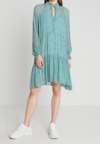 Šaty v barvě modrozelené s tečkovaným vzorem, dlouhými rukávy, volánkovým lemem a výstřihem ve tvaru klíčové dírky, doplněné bílými teniskami.