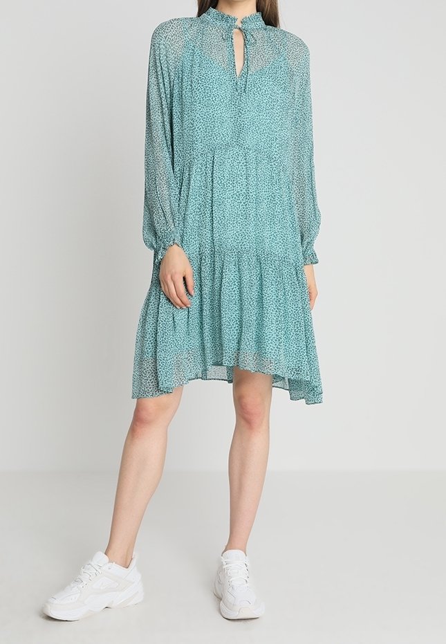 Šaty v barvě modrozelené s tečkovaným vzorem, dlouhými rukávy, volánkovým lemem a výstřihem ve tvaru klíčové dírky, doplněné bílými teniskami.