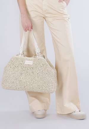 Personne portant un pantalon large crème et des baskets blanches à plateforme, tenant un grand sac tissé beige par les poignées.
