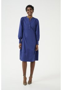 Zila, ceļgalu garuma kleita, kas izgatavota no gludas auduma; tai ir pūkainas piedurknes, izgriezta apkakle un pogu rāvējslēdzējs. Modelis valkā pelēkas augstpapēžu kurpes.