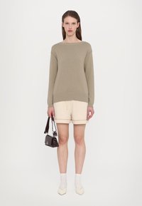 Lys beige strikkegenser med rund hals, kombinert med kremfargede shorts. Holder en liten mørkebrun veske, iført lyse sokker og spisse sko.